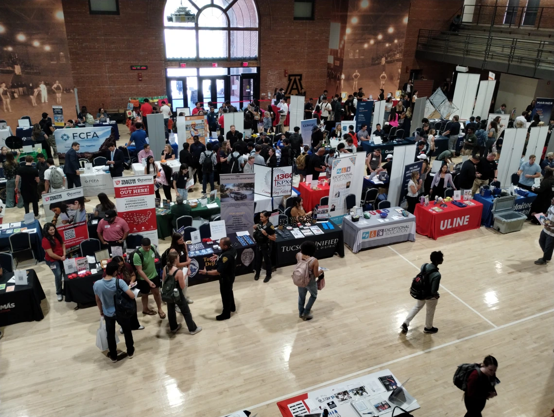 A college fair in a large hall filled with tables and banners and people visiting 